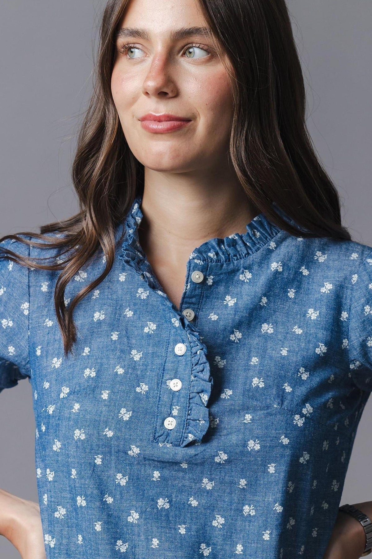 A woman models a blue organic cotton chambray top adorned with small white floral patterns. The shirt features a ruffled collar and buttons, showcasing a casual yet feminine style.