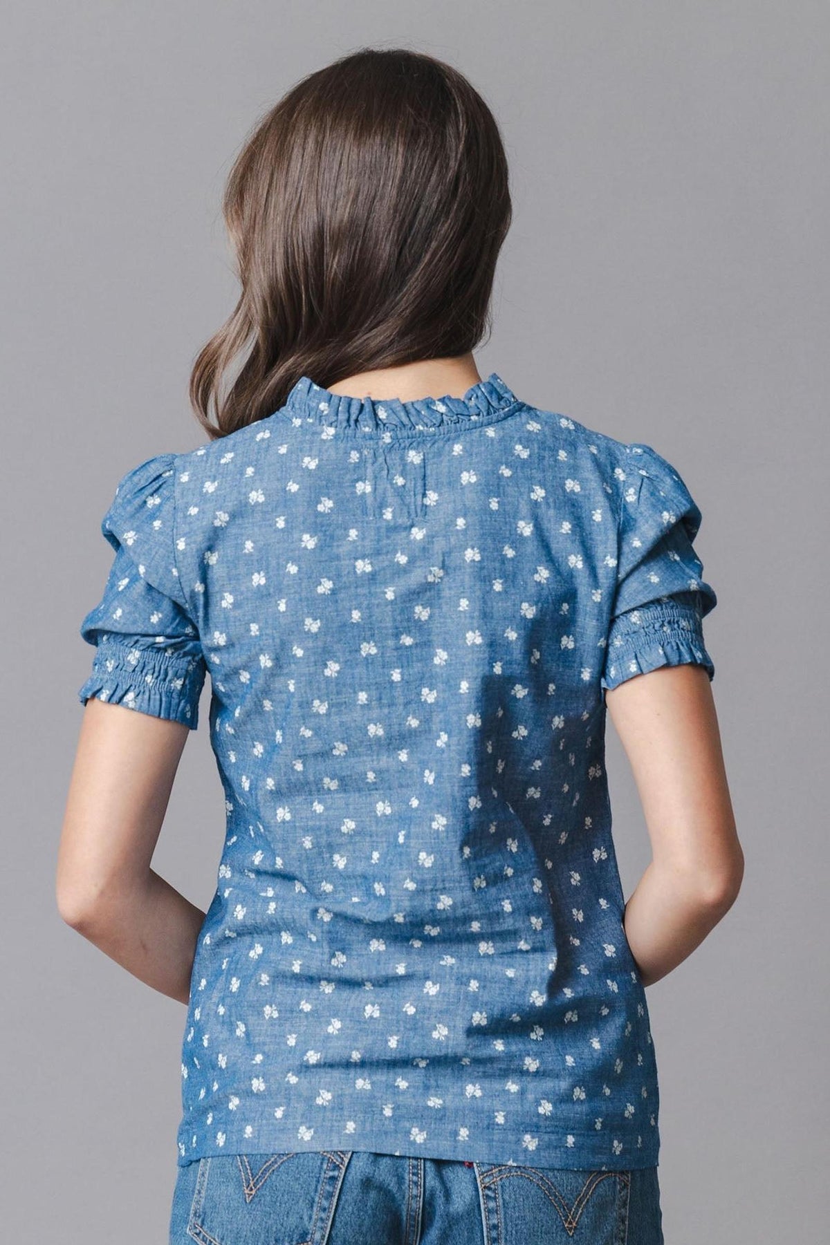 A woman is shown from the back wearing a blue organic cotton chambray top with puff sleeves and a ruffled collar.