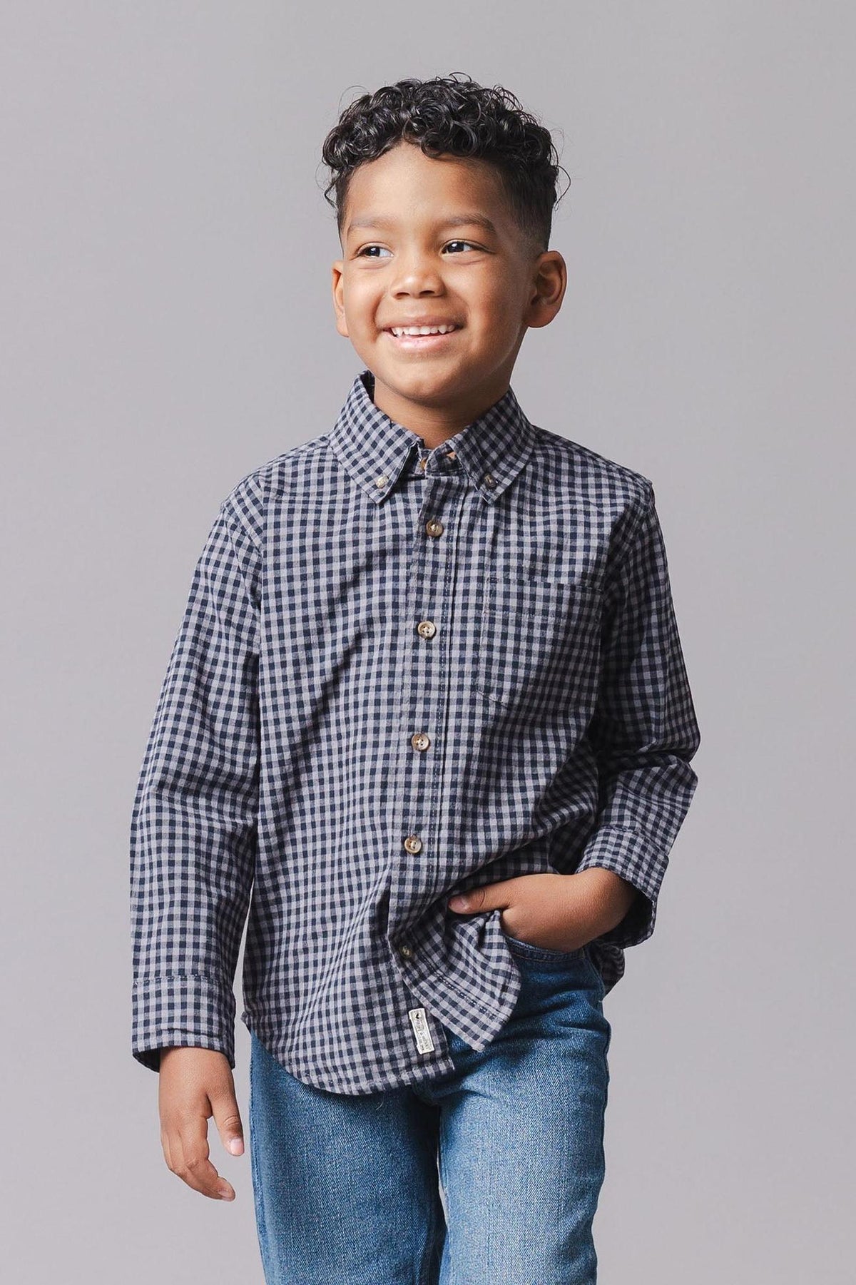 A young boy smiles while wearing a black gingham organic cotton poplin shirt with long sleeves and button-front closure, paired with blue jeans against a neutral background.