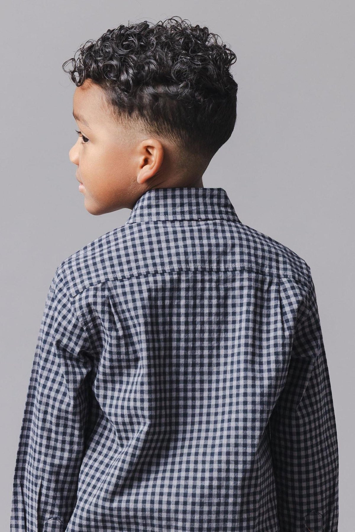 A young boy with curly hair is shown from the back, wearing a black gingham organic cotton shirt.