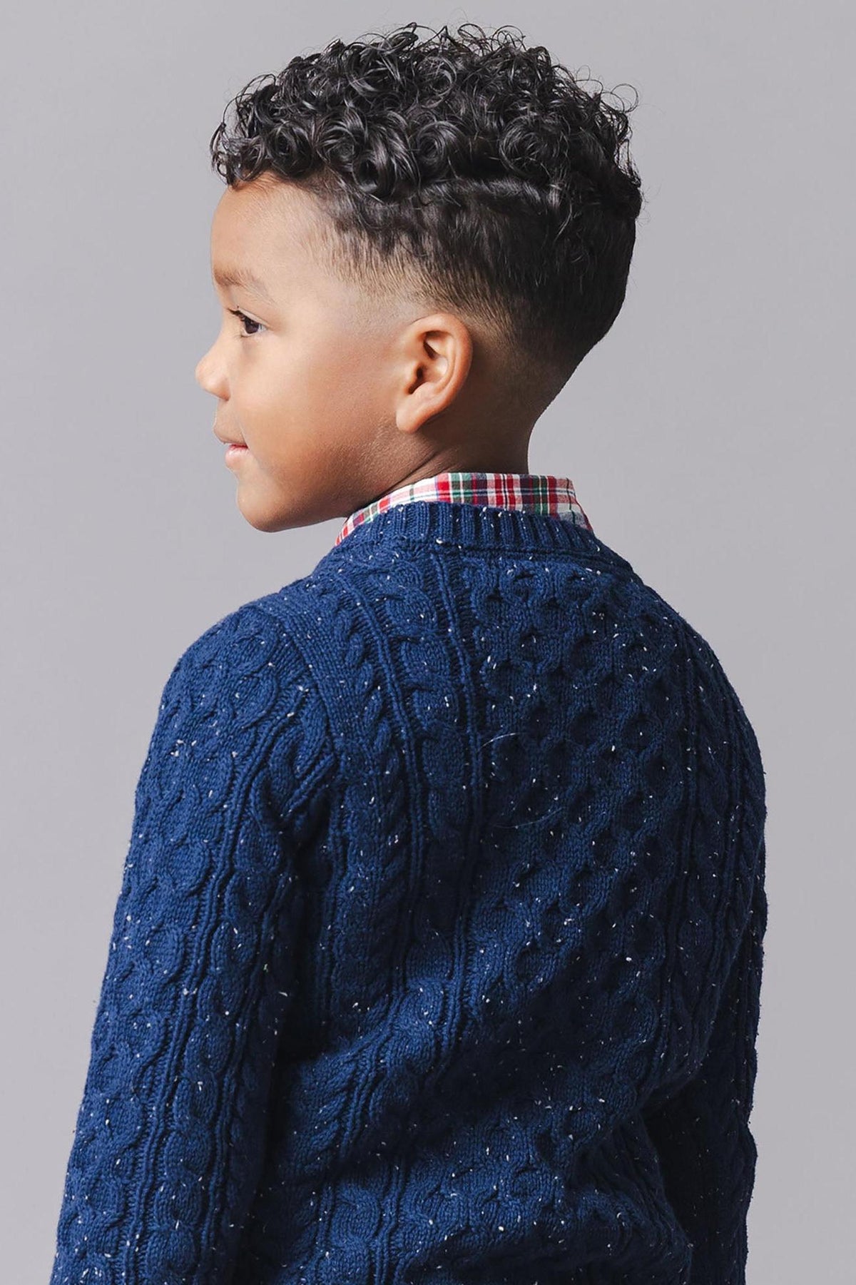 A young boy with curly hair is wearing a navy organic cotton fisherman sweater, featuring a textured cable knit design, paired with a plaid button-down shirt underneath.