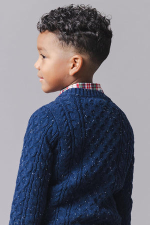 A young boy with curly hair is wearing a navy organic cotton fisherman sweater, featuring a textured cable knit design, paired with a plaid button-down shirt underneath.