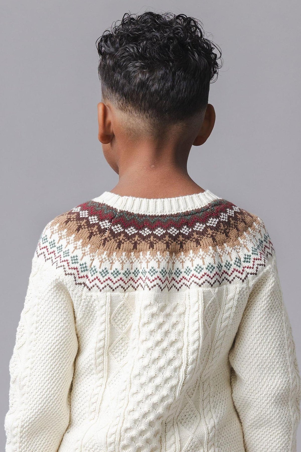 A child’s back is visible, showcasing a cozy ivory sweater with a fair isle design in brown, green, and red. The sweater features a cable texture, made from organic cotton.