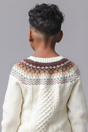 A child’s back is visible, showcasing a cozy ivory sweater with a fair isle design in brown, green, and red. The sweater features a cable texture, made from organic cotton.
