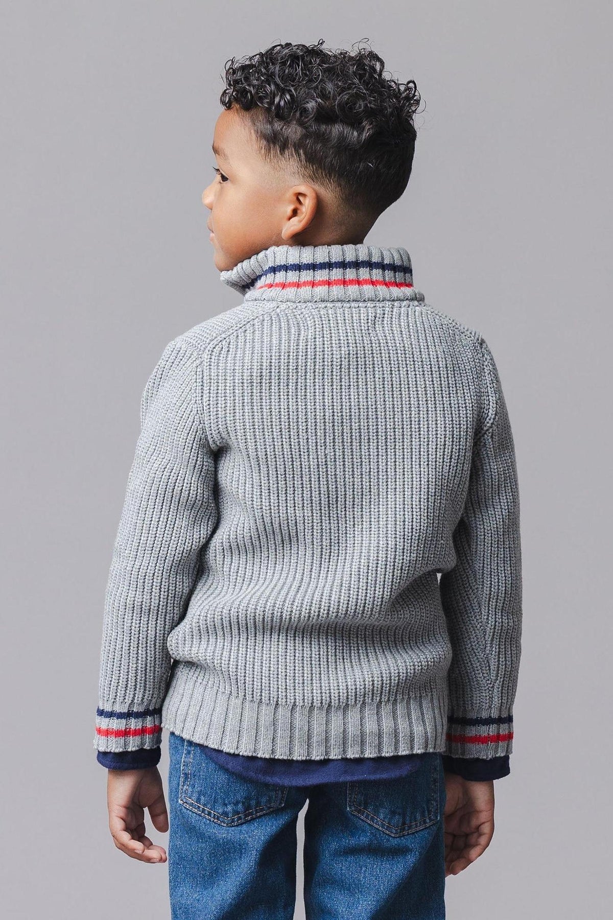 A young child is shown from the back wearing a dark gray organic cotton half-zip sweater with a ribbed texture and red striped details on the collar and sleeves.
