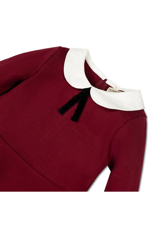 A close-up of a toddler dress in oxblood red featuring a crisp white Peter Pan collar and a black velvet bow. The dress showcases a soft, stretchy ponte knit material.