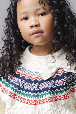 A young girl with curly hair wears an ivory organic cotton sweater featuring a fair isle design and a bow detail at the mock neck, set against a gray background.