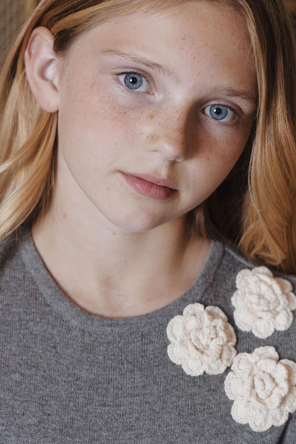 A young girl with blonde hair and freckles wears a dark gray sweater dress featuring three cream-colored rosettes at the neckline, showcasing a soft, organic cotton material.