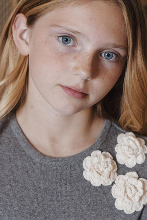 A young girl with blonde hair and freckles wears a dark gray sweater dress featuring three cream-colored rosettes at the neckline, showcasing a soft, organic cotton material.