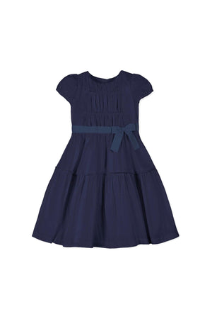 A navy organic cotton tiered party dress featuring puff sleeves and a shirred bodice. It includes a bow detail at the waist, perfect for toddlers and girls.