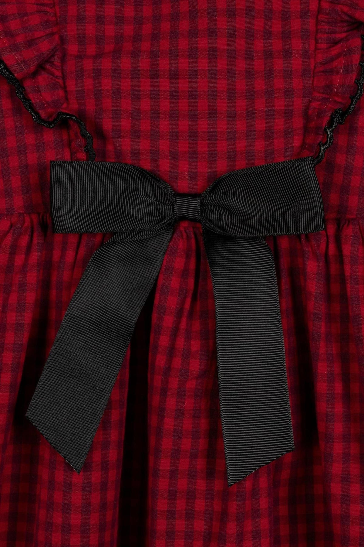 Close-up of a red gingham jumper dress featuring ruffled edges and a black ribbon bow detail at the back.