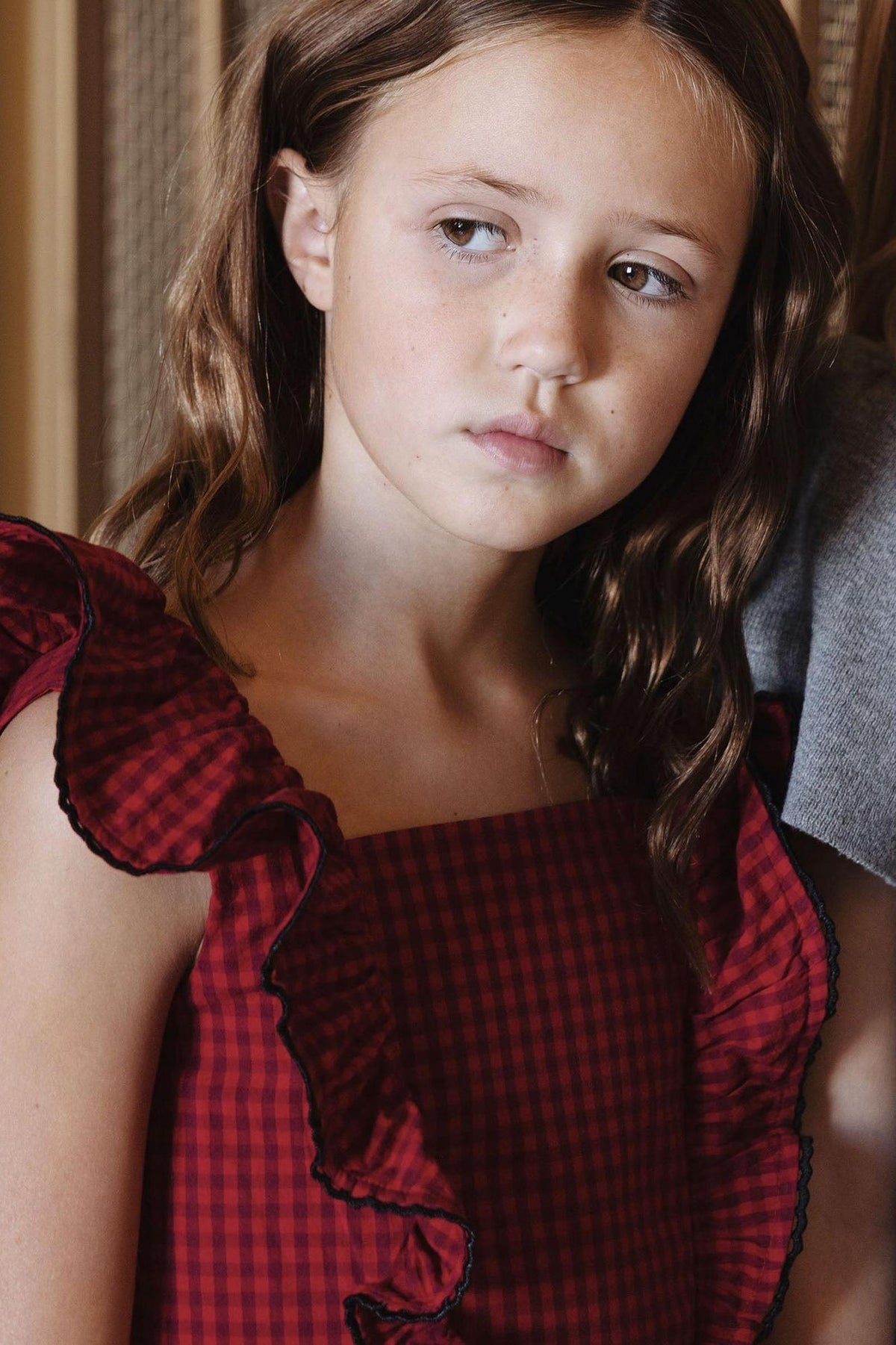 A girl models a red gingham organic ruffle jumper dress with ruffled sleeves and contrasting trim, showcasing a stylish, playful look suitable for toddlers and young girls.