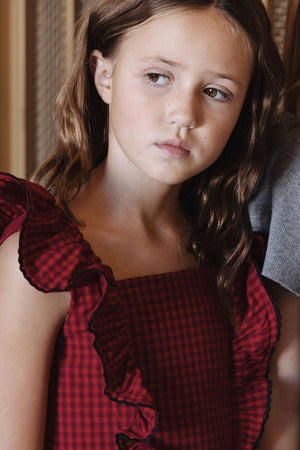 A girl models a red gingham organic ruffle jumper dress with ruffled sleeves and contrasting trim, showcasing a stylish, playful look suitable for toddlers and young girls.
