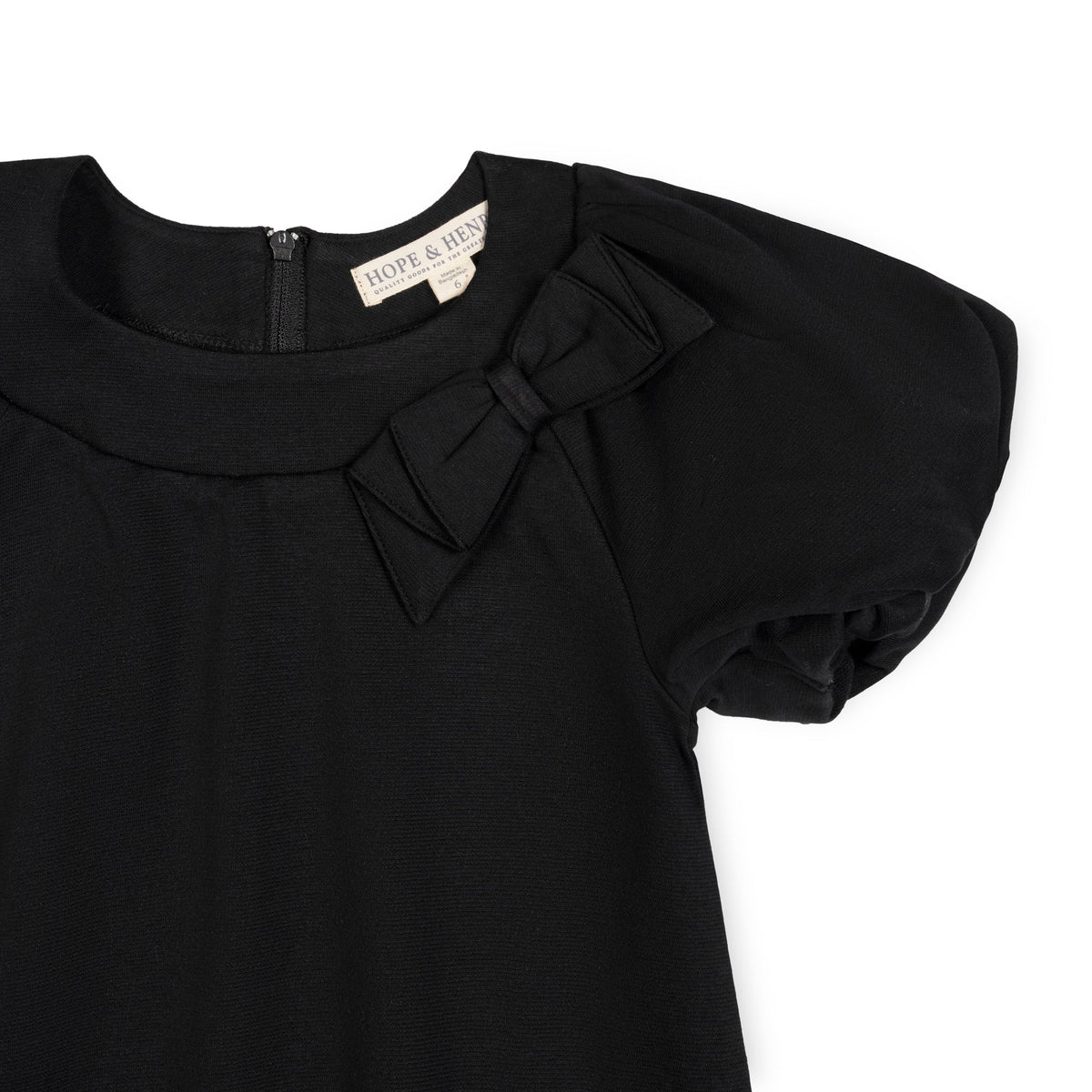 A close-up of a black organic cotton dress featuring bubble sleeves and a bow detail at the neckline, with a zipper closure visible at the back.