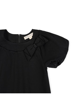 A close-up of a black organic cotton dress featuring bubble sleeves and a bow detail at the neckline, with a zipper closure visible at the back.