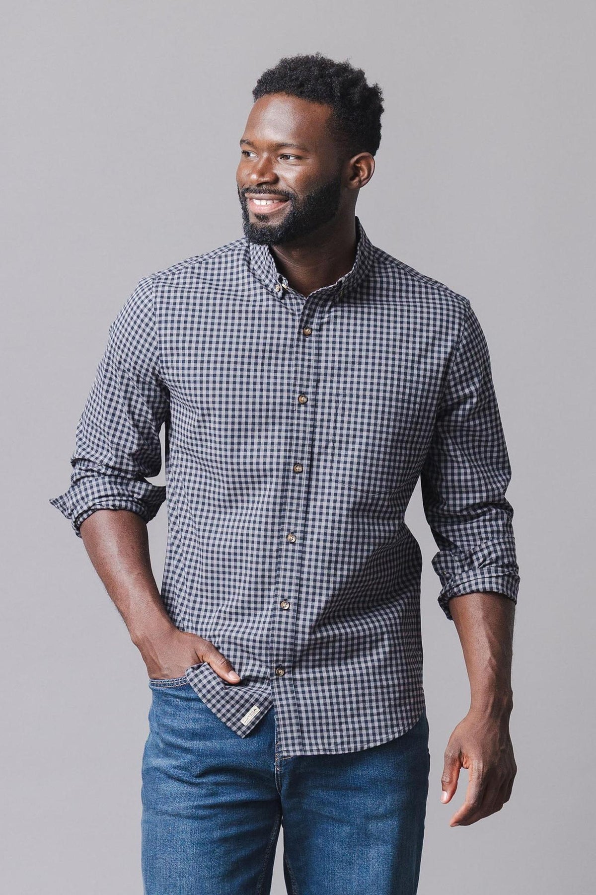 A man smiles while wearing a black gingham organic cotton poplin shirt, featuring a button-down front and a casual style. He pairs it with blue jeans against a neutral background.