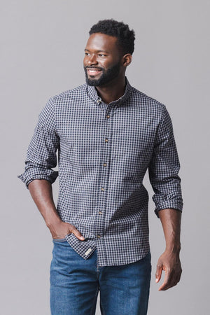A man smiles while wearing a black gingham organic cotton poplin shirt, featuring a button-down front and a casual style. He pairs it with blue jeans against a neutral background.