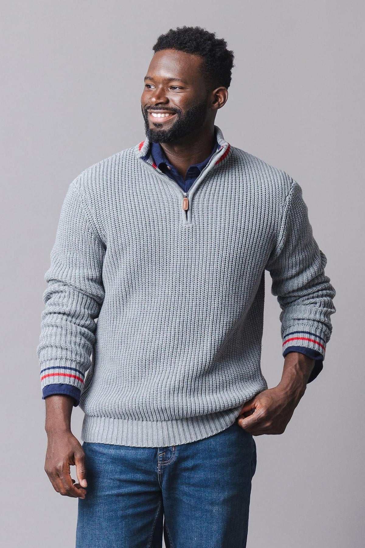 A man smiles while wearing a light gray organic cotton half-zip pullover with a rib knit texture and contrasting red and blue stripes on the cuffs.
