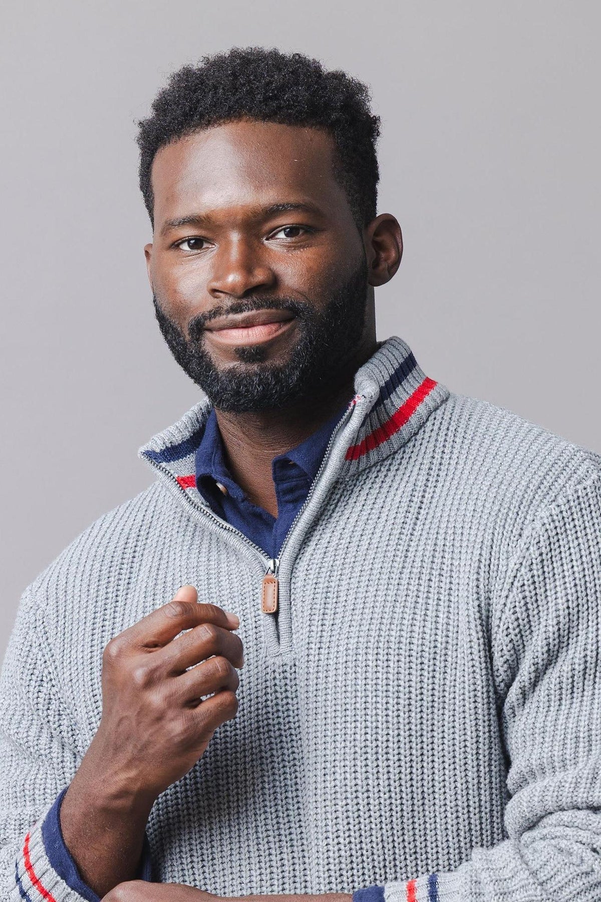 A man wearing a textured gray half-zip pullover with red and blue stripes at the collar stands against a neutral background. The pullover is crafted from soft, sustainable materials.