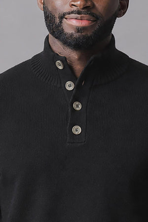 A close-up of a man wearing a black organic mock neck sweater featuring a Henley-style button placket. The fabric appears soft and structured, suitable for casual or polished looks.