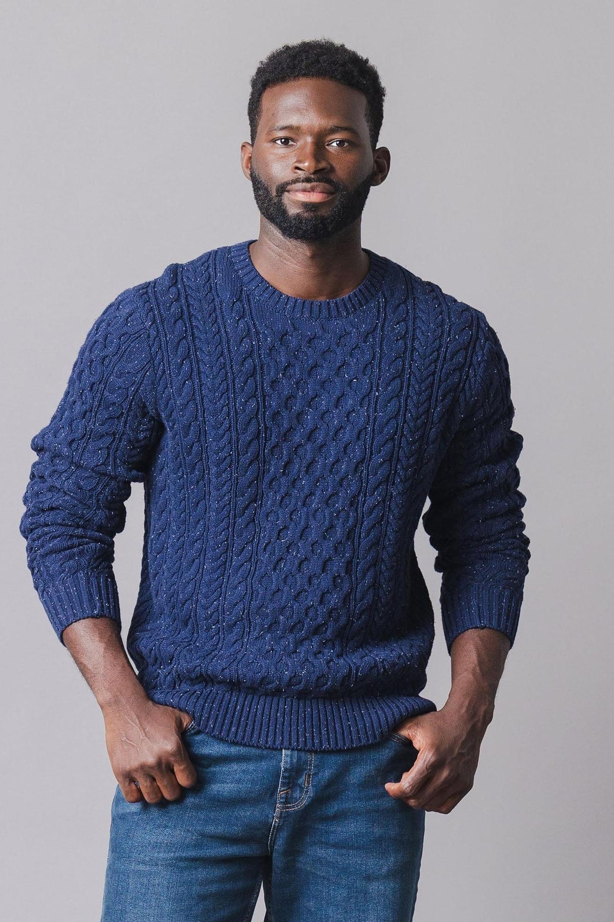 A man stands confidently in a navy organic cotton fisherman sweater featuring an allover cable knit design. The crewneck pullover is both stylish and warm, perfect for cooler seasons.