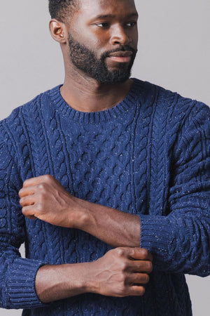 A man wearing a navy organic cotton fisherman sweater with a textured cable knit design stands against a neutral background, showcasing the sweaters cozy and stylish appearance.