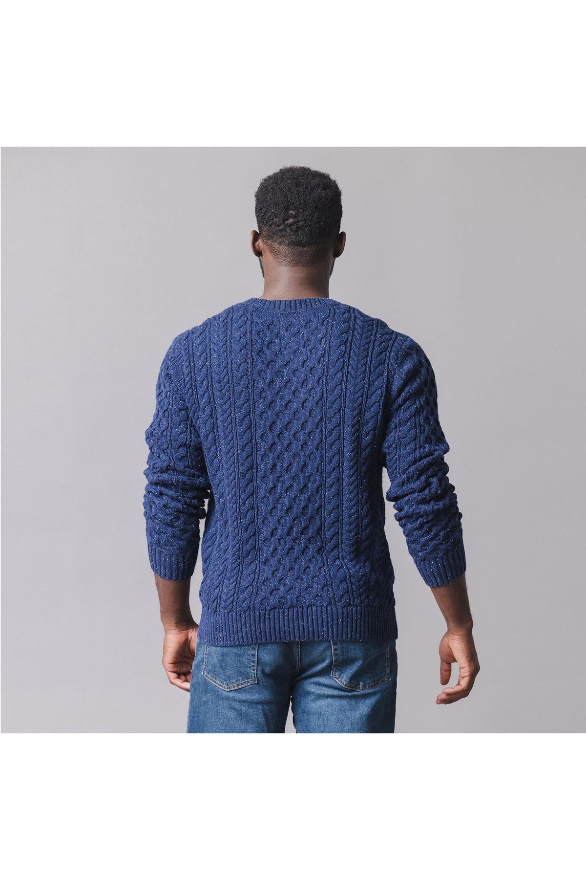 A man is seen from the back wearing a navy organic cotton fisherman sweater featuring an intricate cable knit design.