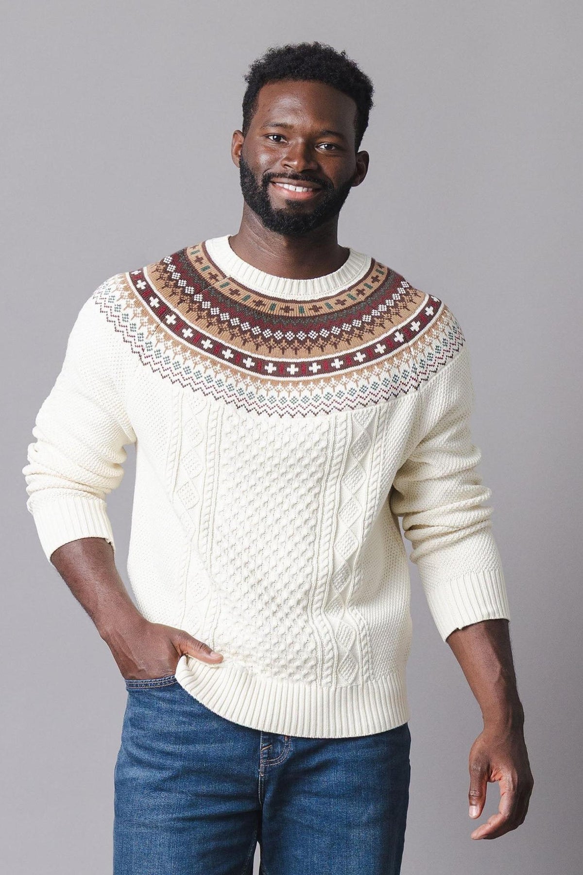 A man models a cozy ivory sweater featuring a fair isle design in brown and red at the shoulders, complemented by a textured cable knit pattern.