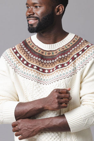 A man smiles while wearing an ivory organic cotton sweater featuring a festive fair isle design in brown and green. The textured cable knit adds warmth and style.