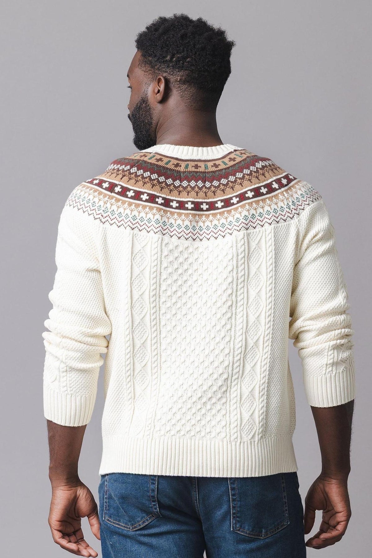 A man is seen from the back wearing an ivory sweater featuring a fair isle design in brown and red at the shoulders, complemented by a textured cable knit pattern.