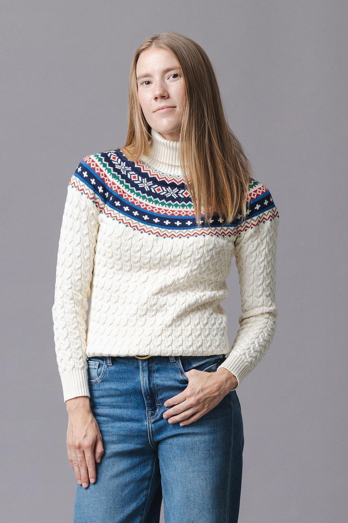 A woman models an ivory organic cotton sweater featuring a fair isle design in red and navy at the shoulders. The cozy knit has a turtleneck and ribbed cuffs.