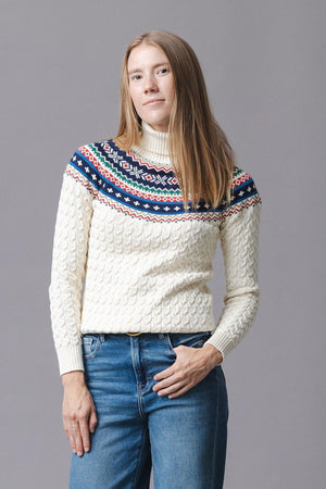 A woman models an ivory organic cotton sweater featuring a fair isle design in red and navy at the shoulders. The cozy knit has a turtleneck and ribbed cuffs.