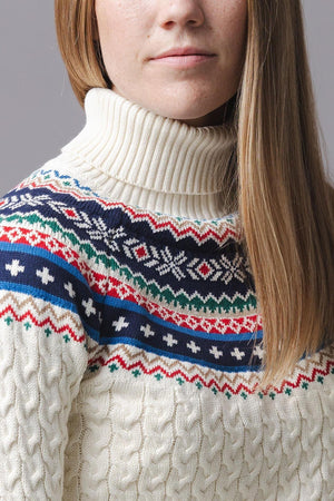 A woman models an ivory organic cotton sweater featuring a fair isle design in red, blue, and green, with a cozy ribbed turtleneck and cable-knit texture, perfect for winter wear.