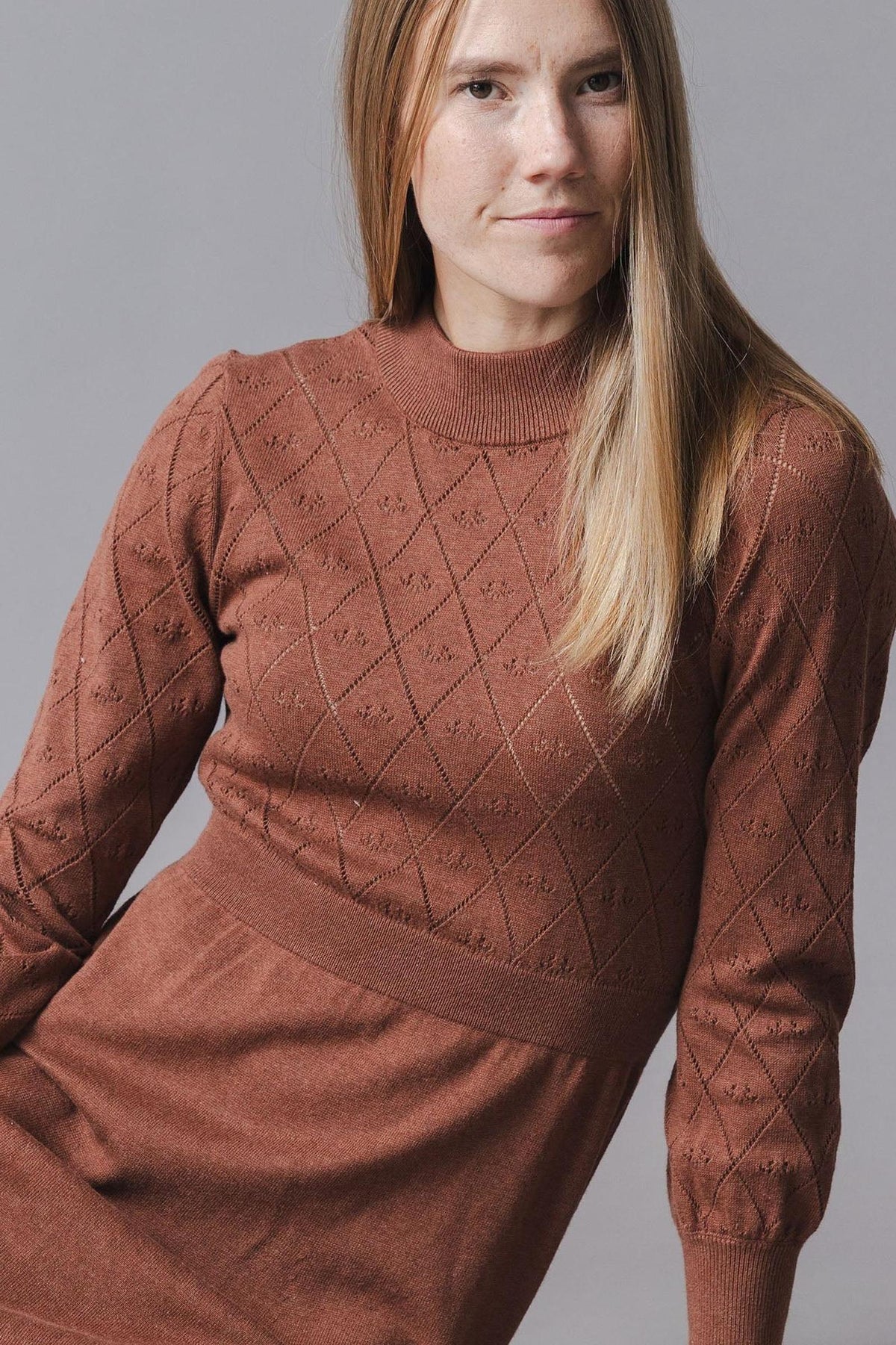 A model wears a cinnamon-colored organic cotton sweater dress featuring a pointelle stitch pattern and puffed sleeves, showcasing a flattering silhouette against a neutral gray background.