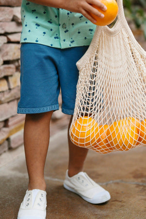 A child wearing stone blue shorts and a light green patterned shirt holds a mesh bag filled with oranges, standing on a stone pathway surrounded by greenery.