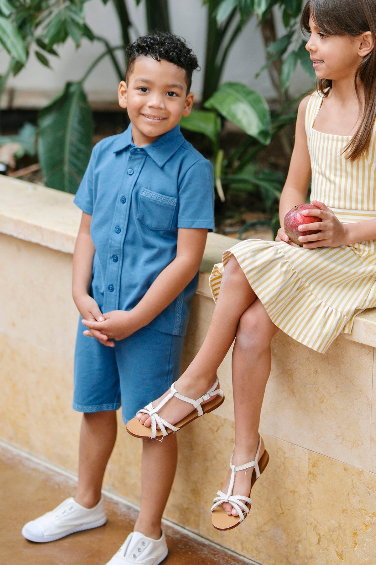 A boy in a blue short-sleeve shirt and matching shorts stands next to a girl in a yellow striped dress holding an apple, surrounded by lush green plants.