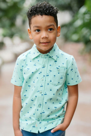 A young boy stands outdoors wearing a short-sleeve, light blue button-down shirt adorned with a playful palm print. The shirt features a classic collar and a front pocket.