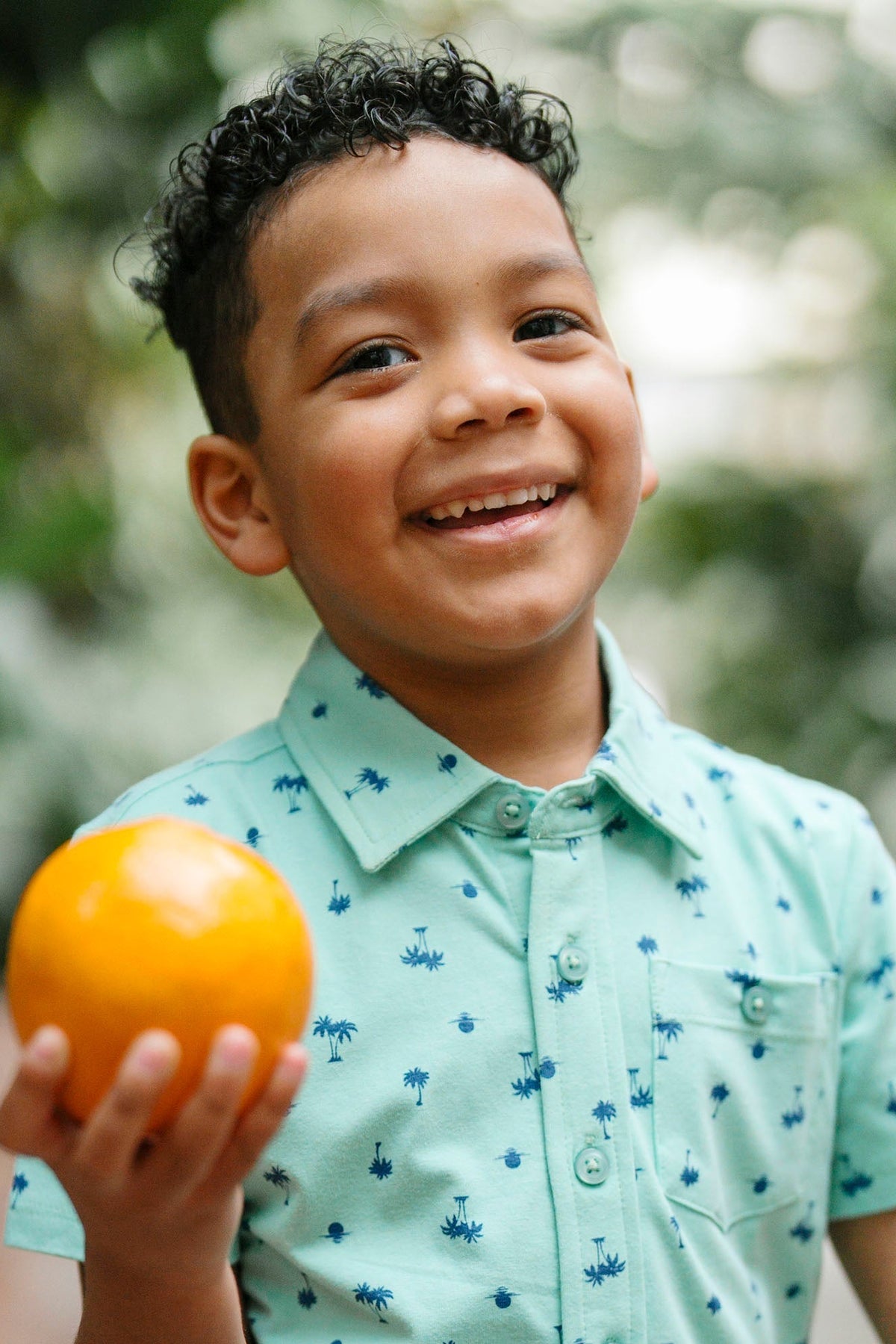 A smiling boy holds an orange while wearing a light green button-down shirt featuring a small palm print. The background is lush with greenery, adding a natural, vibrant feel.