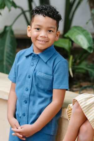 A young boy smiles while wearing a stone blue organic cotton short sleeve button-down shirt with a chest pocket. He stands outdoors, surrounded by lush green plants.