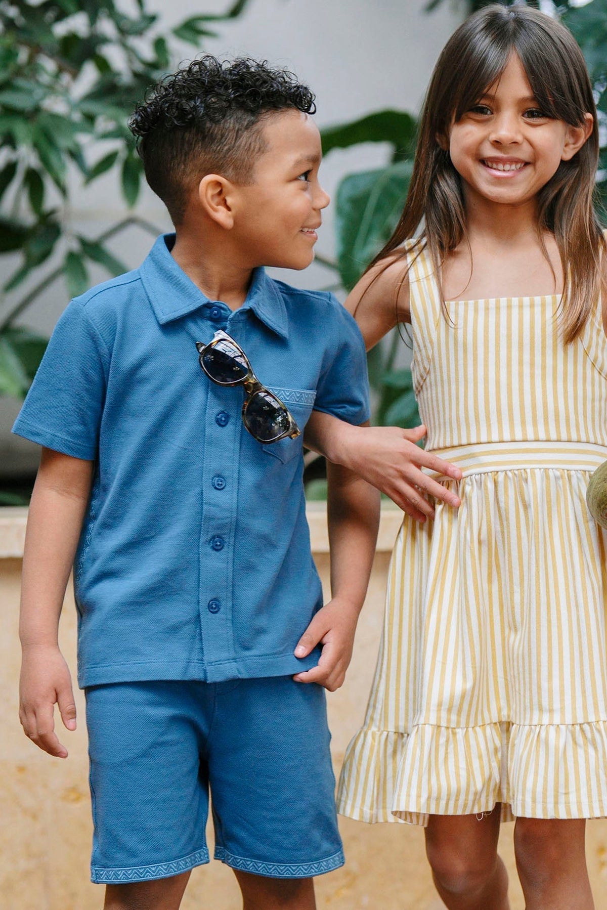 A boy wears a blue short-sleeve button-down shirt with a pocket, while a girl in a yellow striped dress holds a mango, both smiling in a lush garden setting.