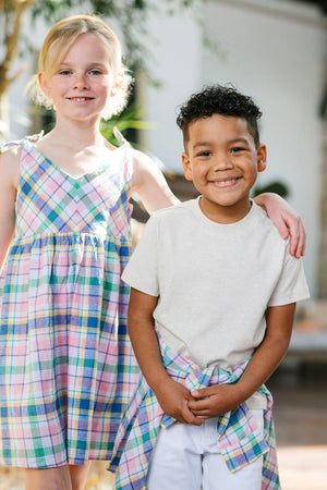 Two children stand together outdoors, smiling. The girl wears a colorful plaid dress, while the boy sports a neutral organic cotton tee and white shorts, showcasing a playful, casual style.