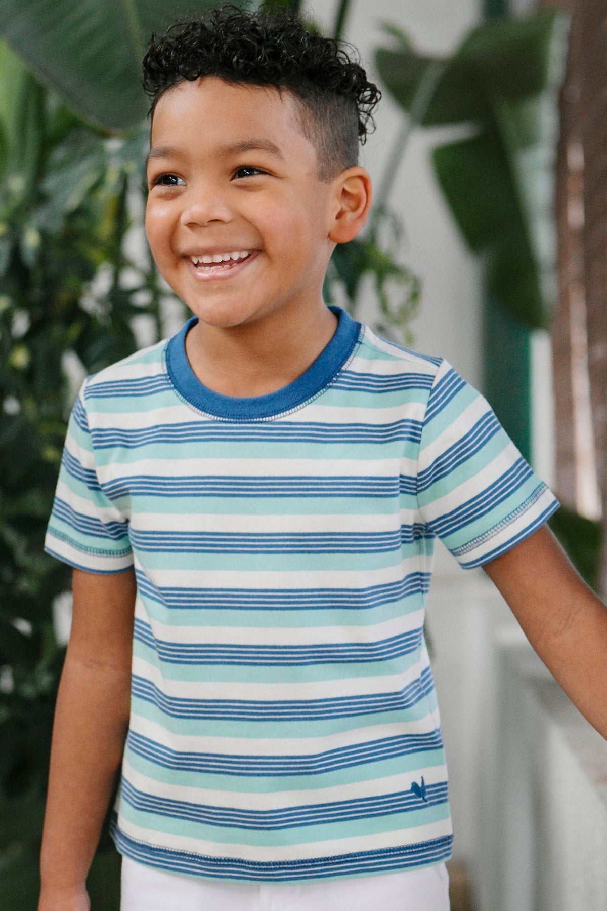 A smiling boy wears a short-sleeve, Tidal Stripe Henley tee made from organic cotton. The shirt features blue and green stripes, offering a vibrant, breathable option for warm weather.