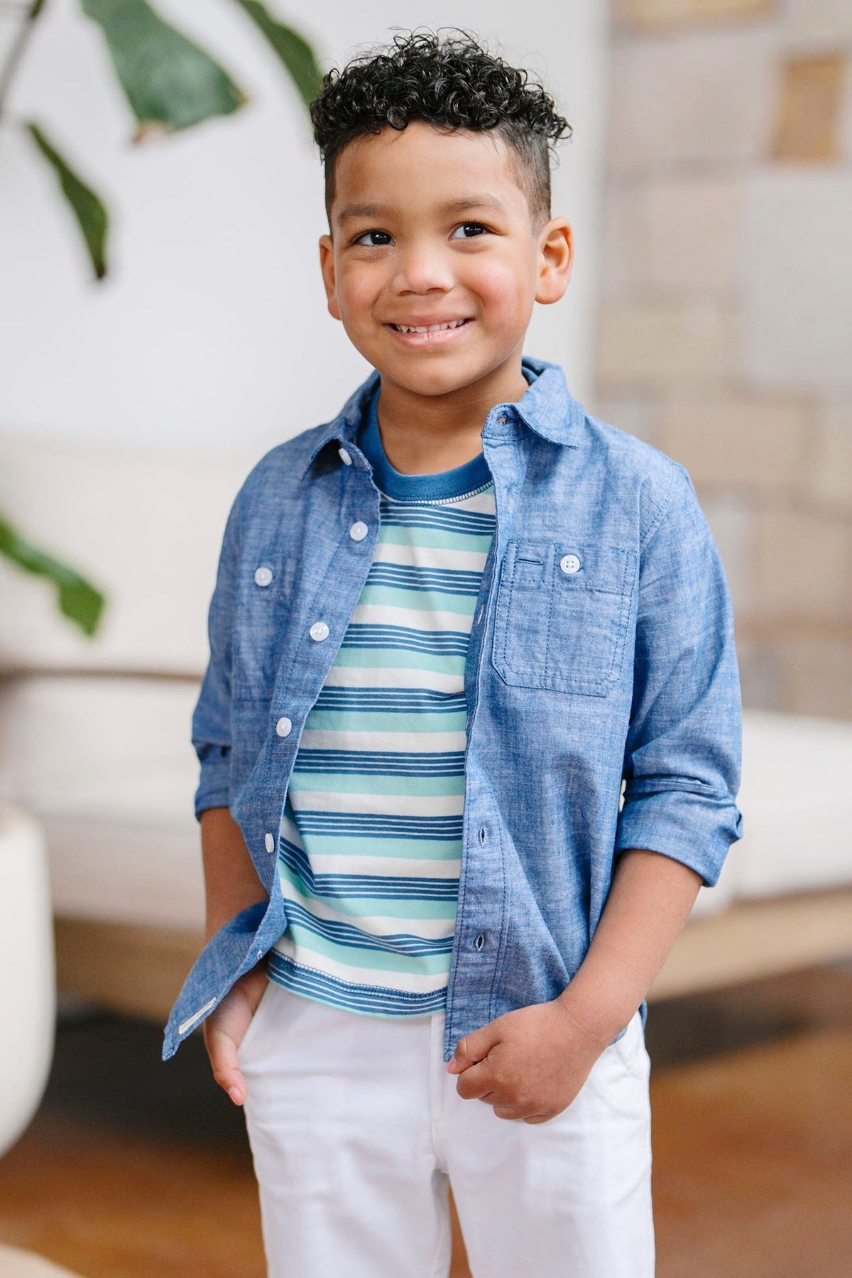 A smiling boy wears a Tidal Stripe organic cotton Henley tee layered under a blue chambray button-up shirt. He stands in a bright.
