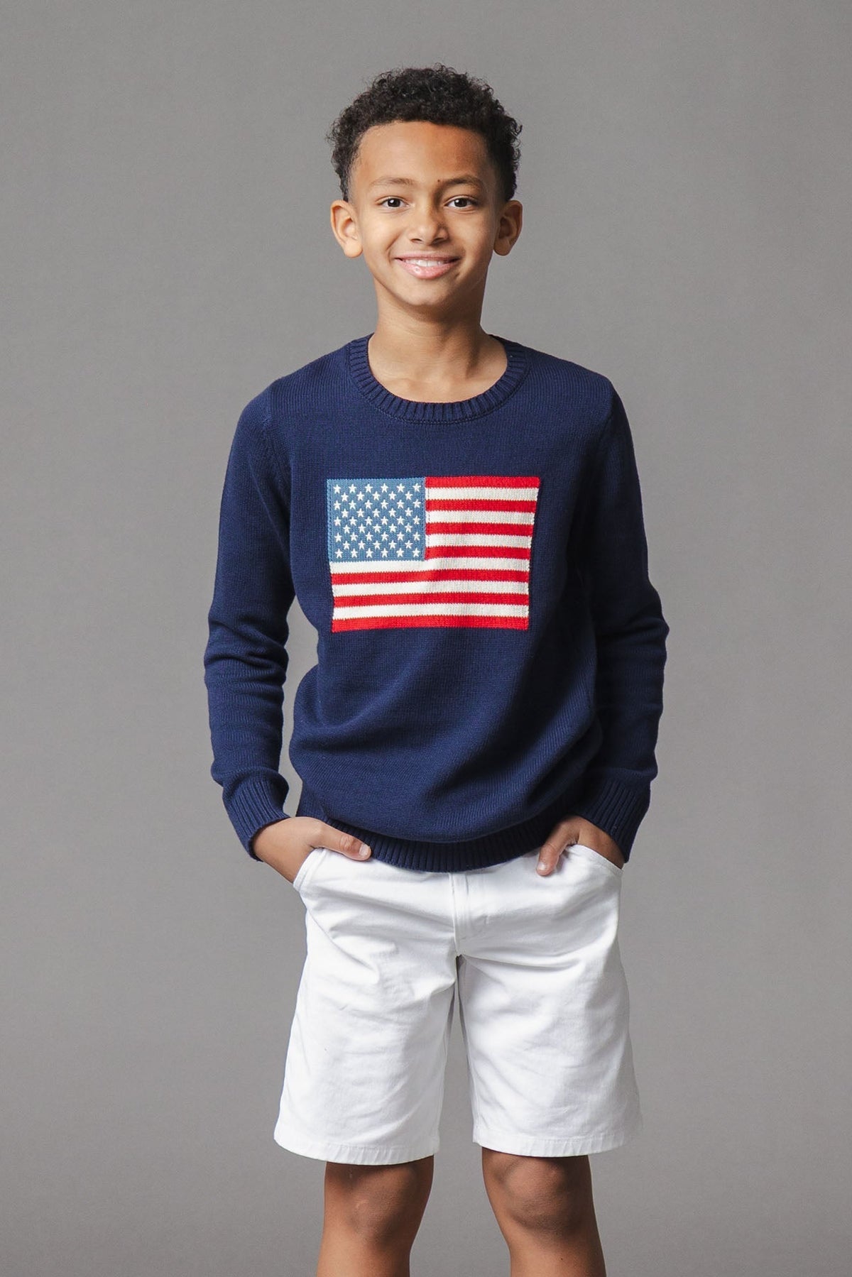 A smiling boy wears a navy blue sweater featuring a bold American flag design on the front, paired with white shorts.