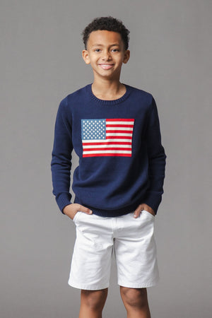 A smiling boy wears a navy blue sweater featuring a bold American flag design on the front, paired with white shorts.