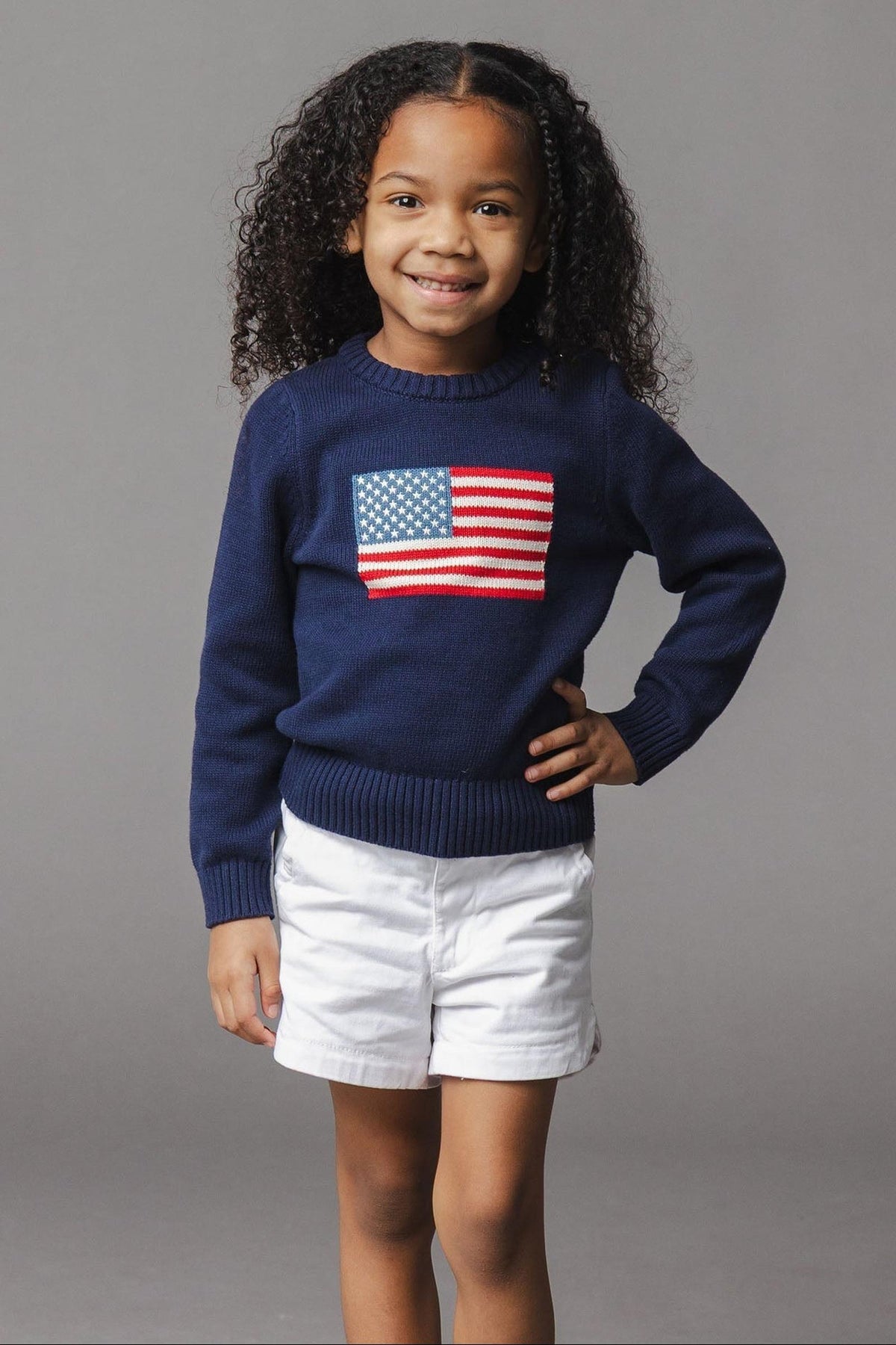 A young girl smiles while wearing a navy organic cotton sweater featuring a flag design on the front. She pairs the sweater with white shorts, standing against a gray backdrop.