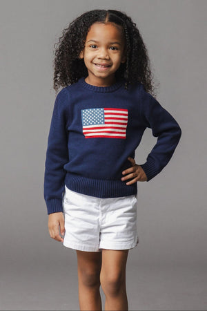 A young girl smiles while wearing a navy organic cotton sweater featuring a flag design on the front. She pairs the sweater with white shorts, standing against a gray backdrop.