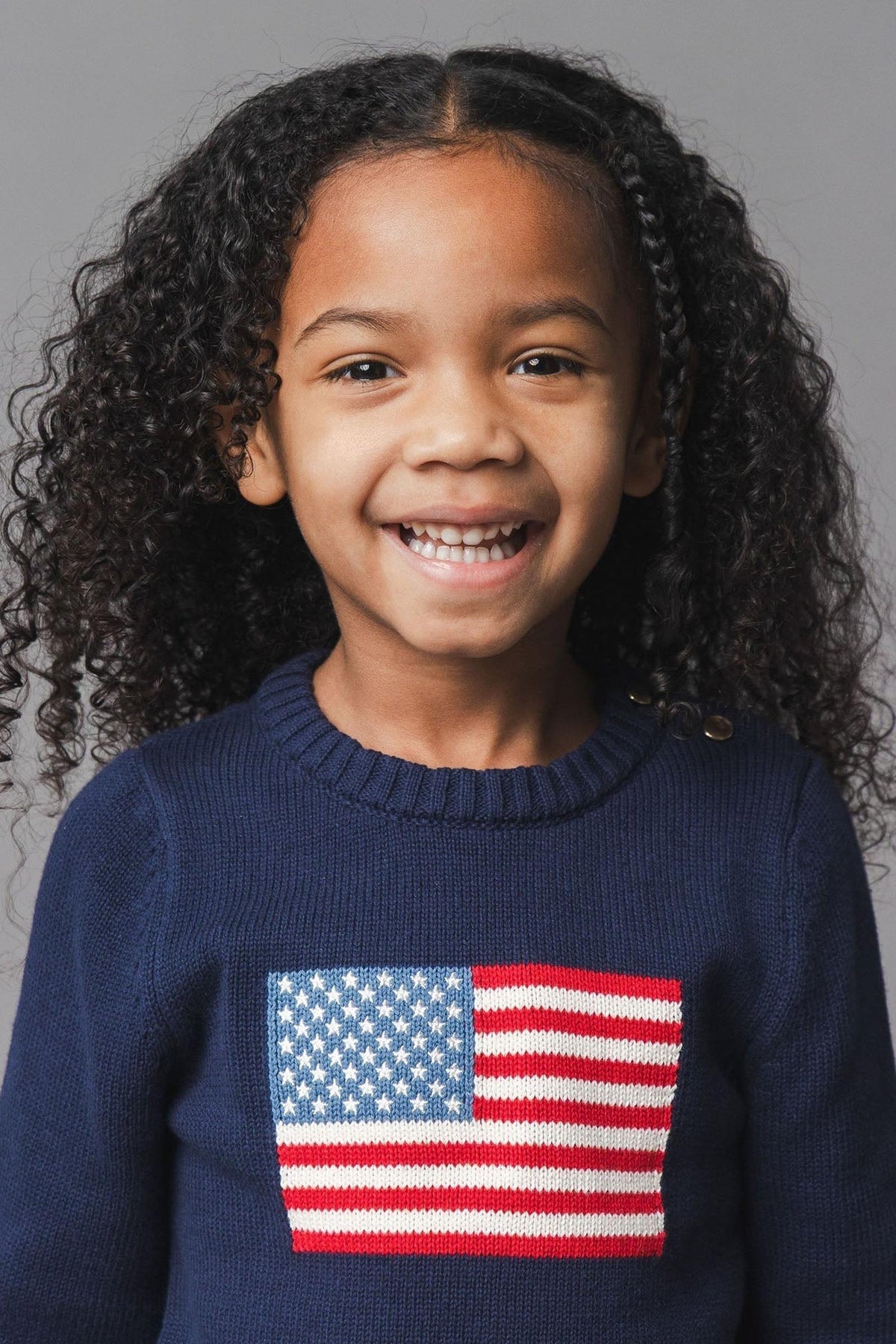 A smiling child with curly hair wears a navy sweater featuring an American flag intarsia design.