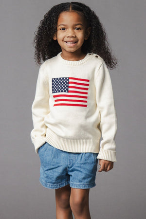 A smiling child wears an ivory organic cotton crew neck sweater featuring a patriotic American flag design. The outfit is complemented by denim shorts, set against a neutral gray background.