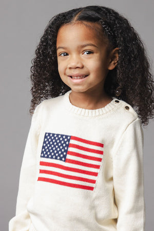 A young girl smiles while wearing an ivory organic cotton sweater featuring a red and blue American flag design on the front.
